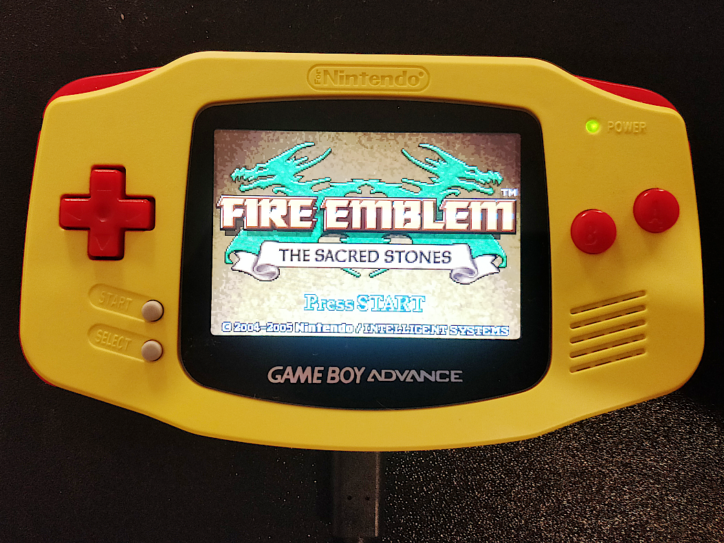 A photograph of a modified Game Boy Advance console. The console is in a yellow shell with red buttons. The screen is backlit and the title screen of the game Fire Emblem: The Sacred Stones can be seen. At the bottom of the photograph a USB-C cable can be seen providing power to the console