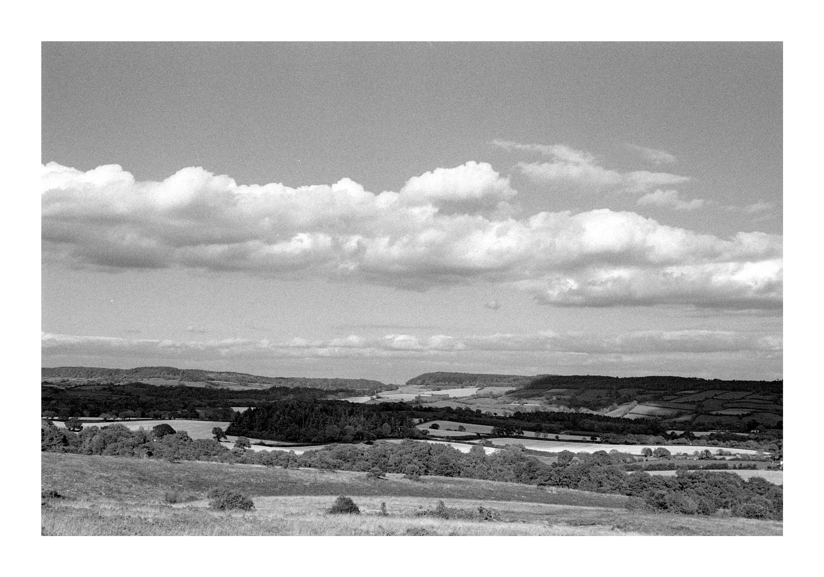 Landscape photograph of moorland, woods and copses. Scatter white clouds are dotted about the sky and cast shadows on the ground.