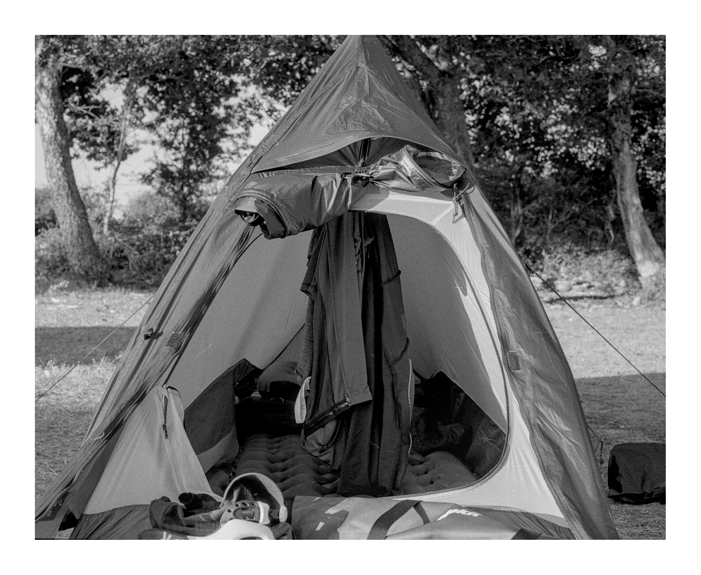 Square cropped photograph. A view into the entrance of a small, one-person tent. Sleeping equipment is visible inside, as well as cycling clothes hanging from a hook.
