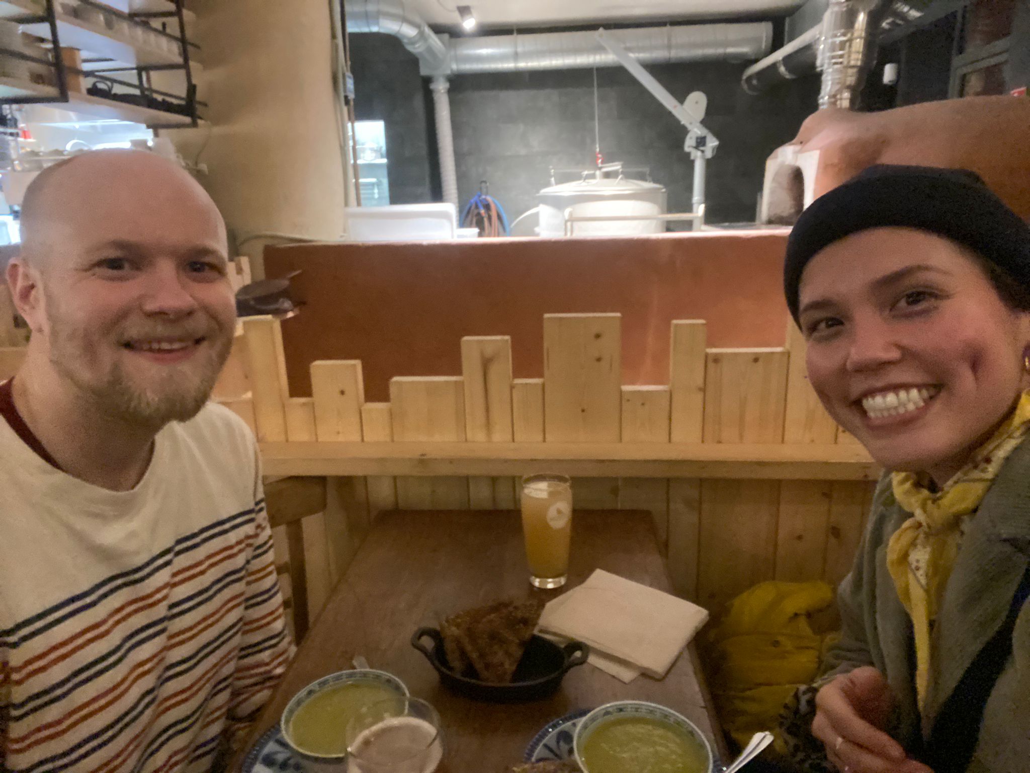 Two people sat in a restaurant taking a self-portrait. On the table is fresh, handmade bread, soup and glasses of beer and cola. On the left is a man in his 30s with very short blonde hair, a beard and a light coloured top with blue-and-orange, horizontal stripes. On the right is a young woman, who is taking the picture, wearing a green coat, black hat and yellow neck-scarf. In the rear of the room, is a large pizza oven and brewing equipment