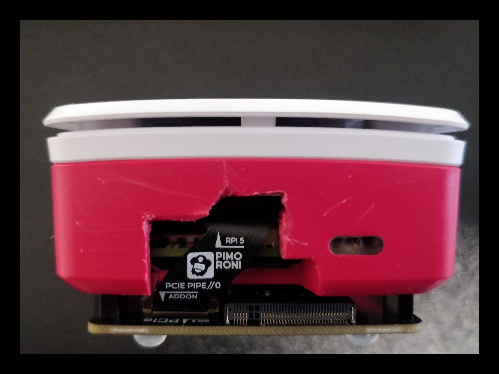 An end-on view of a Raspberry Pi 5 case. An addon board is attached to the bottom of the case. A clumsy hole has been cut into the case, to allow a ribbon cable to run from the addon board to the Raspberry Pi inside the case. Tool marks are visible around the edge of the hole.
